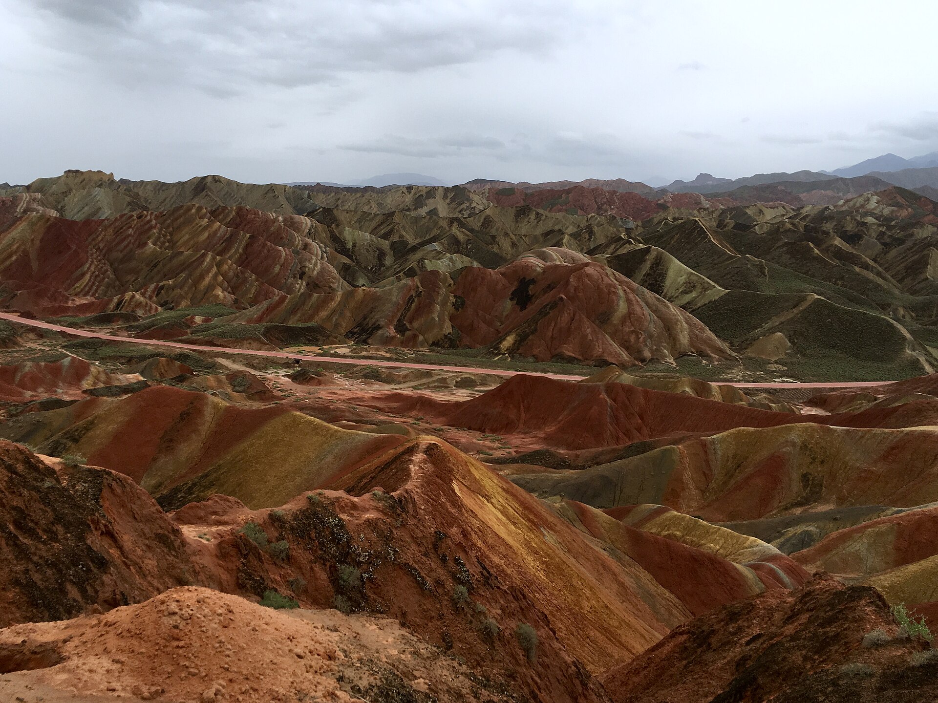 张掖丹霞的深红与绿色丘陵位于灰色雨云下，一条道路横过谷地。